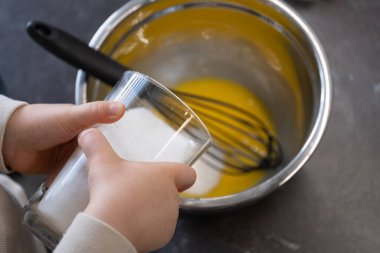 İçinde yumurta ve çırpı olan karıştırma kabının yanına yerleştirilmiş şekerle dolu bir bardağı tutan küçük çocukların yakın plan fotoğrafları. Bu samimi detay erken mutfak müdahalesini vurguluyor, duyusal öğrenme için ideal, çocuklarla yemek pişirmek,