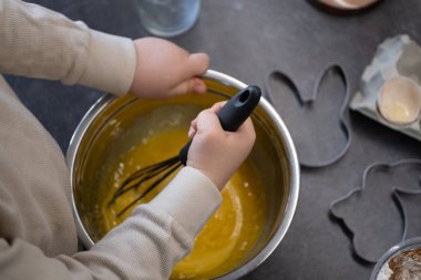 Kurabiye keskisi ve unla çevrili metal bir kasede hamuru karıştıran çocuk ellerine yakın plan. Paskalya tavşanı ve ayı şekilli kesiciler tatil pişirmesini işaret ediyor. Duyusal oyun, çocuklarla yemek pişirme, Paskalya hazırlığı ve ebeveynlik blogları için harika.