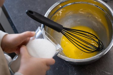 Küçük bir çocuk içinde çırpılmış yumurta sarısı ve çırpılmış çırpılmış çırpılmış metal bir kasenin üzerinde bir bardak şeker tutuyor. Yakın çekim, çocukların yemek pişirme, erken duyusal gelişim ve aşçılık eğitimi için uygun bir bakış açısını yakalar.