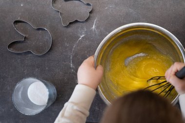 Sarı hamuru, şeker ve fırın kalıplarıyla çevrili metal bir kasede çırpıcıyla karıştıran bir çocuğun yukarıdan görünüşü. Çocuklarla, tariflerle ya da yemek bloglarında görselleştirmeler yapmak için mükemmel..
