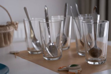 Metal kaşıklı şeffaf bardaklar, Paskalya yumurtası boyamaya hazır bir masanın üzerinde. Şenlik düzeninde kırık bir kurabiye ve sepet ipucu. Tatil, aşçılık hazırlık ve mevsimlik içerik için ideal.