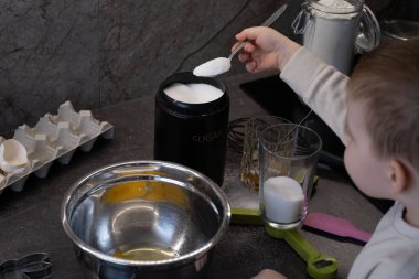 Küçük çocuk evde yemek pişirirken karıştırma kabına bir kaşık şeker ekliyor. Sahnede tezgahta şeker, yumurta ve mutfak aletleri yer alıyor. Bu fotoğraf gerçek bir öğrenme anını yakalıyor.