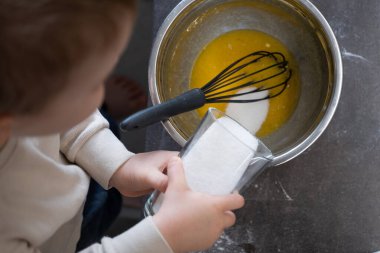 Küçük çocuk çırpılmış yumurtalarla bir kaseye şeker ekler, bardağı dikkatlice tutar. Çocuk pişirme sürecine tamamen dahil olmuş, erken dönem mutfak becerileri ve duyusal keşifler sergiliyor. Çocuk gelişimi, ev yemeği veya