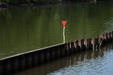Sakin yeşil bir nehirde ahşap bir bariyerde kırmızı üçgen navigasyon işareti. Seyahat, tekne güvenliği, nehir yönetimi ya da minimalist doğa temaları için ideal. Yüksek kalite fotoğraf
