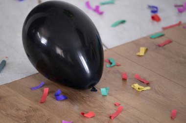 Siyah bir lateks balon, renkli konfetilerle çevrili ahşap ve fayanslı bir zeminde uzanıyor. Sahne kapalı alandaki şenlikli bir kutlamanın sonrasını yakalıyor, doğum günü temalarında, parti dekorasyonlarında, etkinlik pazarlamalarında ve şenlik için mükemmel hale getiriyor.