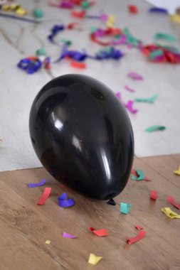 Ahşap ve fayanslı zeminin kenarındaki renkli konfeti ve kıvrımlı kurdelelerle çevrili siyah bir balonun dikey görüntüsü. Çekim bir partiden sonra şenlikli kaosu yakalıyor, kutlama kartları, doğum günü tasarımları, sosyal medya ve hatta