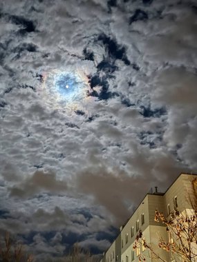 Ay çarpıcı bulutlarla dolu desenli bir gece gökyüzünde parlıyor ve büyüleyici bir halo etkisi yaratıyor. Aşağıda, aydınlık pencereli modern bir bina yukarıdaki gökyüzü manzarasıyla çelişiyor. Ay ışığının ürkütücü parıltısı ve bulutlar...