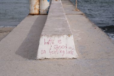 Yıpranmış beton bir iskeleye yakın çekim. El yazısıyla yazılmış grafiti ile hayat güzel. Yani aşkı hisset. Deniz dalgaları arka plana çarparak, insan ifadesiyle doğal çevrenin kontrastını da ekliyor. Yaşam tarzı makalelerinde kullanım için harika.