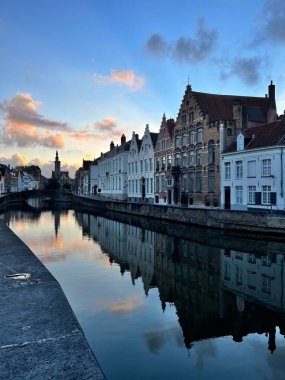 Bruges 'deki kanal kenarındaki büyüleyici binalar altın saat boyunca durgun suya mükemmel bir şekilde yansıyor. Yumuşak günbatımı gökyüzü ve klasik Flaman mimarisi seyahat, mimari ve Avrupa şehir manzarası temaları için ideal barışçıl, resimsi bir sahne yaratır.