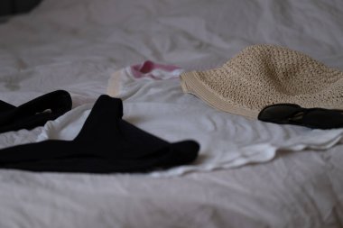 Yaz seyahati eşyalarının minimalist düzlüğü: hasır şapka, güneş gözlüğü, mayo ve yatağa serilmiş bir atlet. Tatil hazırlığı, tatil hayatı, seyahat denetim listesi veya moda içeriği ile ilgili görseller için ideal.