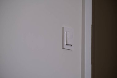 Temiz bir duvarda beyaz, modern bir ışık düğmesi var. Kapalı mekan ışıklarıyla yakalanmış. Akıllı enerji kullanımı, asgari ev tasarımı ve günlük rutinleri temsil eder. Enerji tasarrufu konuları, ev otomasyonu veya kullanım içeriği için uygun.