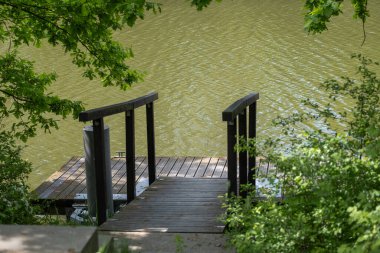 Ahşap bir rıhtım, yeşil yapraklar ve yapraklı dallarla çevrili sakin bir nehre açılıyor. Yaz rahatlama, doğa kaçamakları, balıkçılık, eko-turizm veya farkındalık kavramları için mükemmel bir imaj..