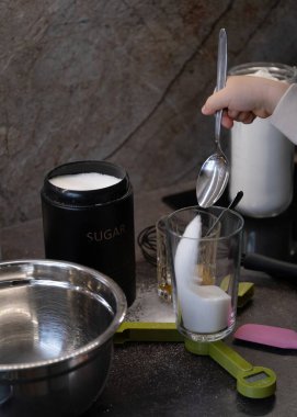 Dijital mutfak terazisinde kaşıkla şeker kepçesiyle şeker kepçeleyen bir çocuğun küçük ellerini kapatın. Sahne un, şeker, yumurta ve mutfak aletleri gibi malzemelerle ev yapımı pişirme sürecini gösteriyor. Eğitim içeriği ve ebeveynlik blogları için ideal, 