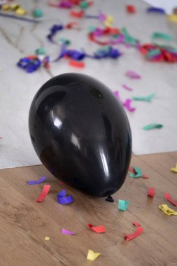 Ahşap ve fayanslı zeminin kenarındaki renkli konfeti ve kıvrımlı kurdelelerle çevrili siyah bir balonun dikey görüntüsü. Çekim bir partiden sonra şenlikli kaosu yakalıyor, kutlama kartları, doğum günü tasarımları, sosyal medya ve hatta