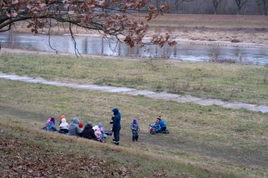Soğuk bir günde, bir nehir kenarındaki çimenli bir arazide oturan bir grup çocuk ve yetişkin. Grup sıcak bir şekilde giyinmiş, doğa içinde bir açık hava aktivitesi veya eğitim oturumunun tadını çıkarmaktadır. Yerleşim, doğal çevresi olan bir topluluğun toplanmasını yakalıyor.