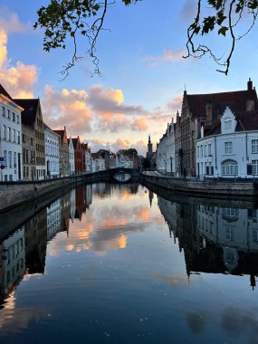 Belçika 'nın Bruges kentindeki bir kanal boyunca uzanan tarihi binaların mükemmel bir yansıması. Yumuşak bulutlar ve pastel gökyüzü bu UNESCO listesindeki ortaçağ şehrinin romantik ve mimari güzelliğini artırıyor. Seyahat ve simetri için ideal.