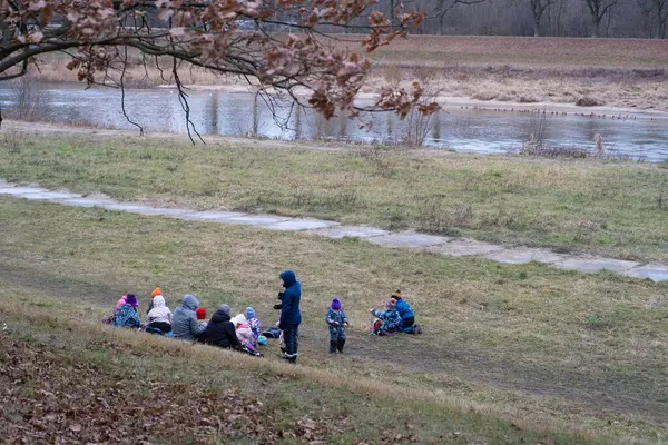 Soğuk bir günde, bir nehir kenarındaki çimenli bir arazide oturan bir grup çocuk ve yetişkin. Grup sıcak bir şekilde giyinmiş, doğa içinde bir açık hava aktivitesi veya eğitim oturumunun tadını çıkarmaktadır. Yerleşim, doğal çevresi olan bir topluluğun toplanmasını yakalıyor.