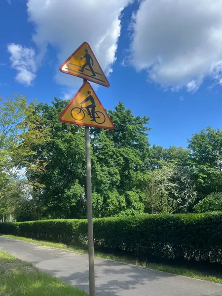 Önümüzde yaya ve bisiklet geçişleriyle ilgili iki sarı üçgen trafik işareti var. Yeşil bir çit ve ağaçların kurduğu bir direğe monte edilmiş. Trafik güvenliği ve kentsel tasarım temaları için uygun