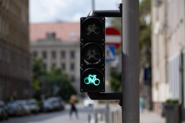 Bulanık bir şehrin arka planında parlayan yeşil bir bisiklet trafik ışığı. Sinyal açıkça görülüyor, bu da bisikletçilerin ilerlemesi için izin verildiğini gösteriyor. Sürdürülebilir ulaşım, şehir bisikleti ve kentsel altyapı gibi konular için kullanışlı.