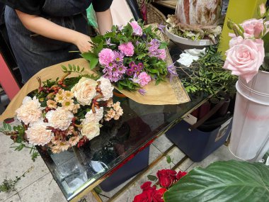 Çiçekçi ellerinin bir çiçekçide iki canlı çiçek buketini sardığı yakın çekim görüntüsü. Bir bukette pembe ve mor çiçekler yer alırken, diğerinde pastel güller ve karanfiller yer alıyor. İdeal