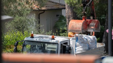 Kazı kovasının büyük beyaz bir çuvalı kargo kamyonuna taşıdığı bir inşaat sahnesinin yakından görüntüsü. Ağaçlar ve konut binaları ile çevrili bu görüntü uygun