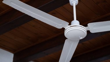 Beyaz tavan vantilatörünün yakın çekim görüntüsü görünür siyah kirişli ahşap bir tavana monte edilmiş. Bu görüntü iç tasarım, yaz konforu, havalandırma ve evler için çevre dostu soğutma kavramlarına uygundur.