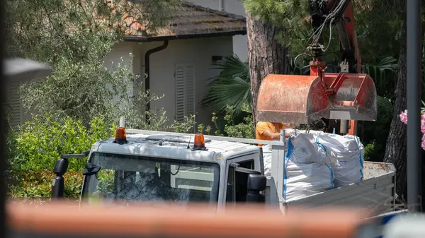 Kazı kovasının büyük beyaz bir çuvalı kargo kamyonuna taşıdığı bir inşaat sahnesinin yakından görüntüsü. Ağaçlar ve konut binaları ile çevrili bu görüntü uygun
