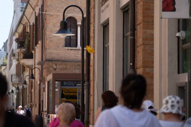 Bir grup insan, tuğla binalar ve dükkan önleriyle dolu eski bir caddede geziniyor. Bir sokak lambasına tek bir sarı yapay çiçek iliştirilerek tuhaf bir dokunuş eklenir. İdeal