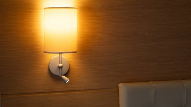 Aydınlatılmış, bej tonlu bir yatak başı lambası, bir otel odasındaki yastıklı yatak başlığının üstündeki ahşap duvara sıcak ışık tutuyor. Konukseverlik için ideal görsel, iç tasarım, rahat yatak odası dekorasyonu ve konaklama promosyonları