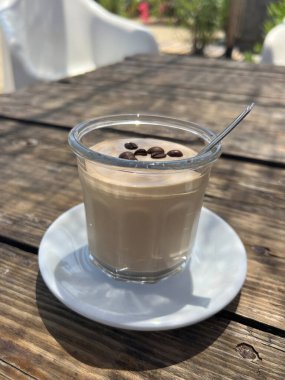 Soğuk kahve köpüğü, bembeyaz bir çay tabağının üzerinde servis edilen cam bardakta metal kaşıkla servis edilir ve çikolata incileriyle süslenir. Doğal güneş ışığı ve yumuşak arka plan bulanıklığı olan kırsal ahşap bir masada yakalanmış. Özetlemek için mükemmel