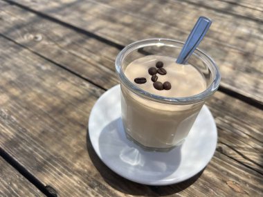 Kremalı soğuk kahve köpüğüyle dolu cam bir fincan, üzerinde kavrulmuş kahve çekirdekleri ve bir kaşıkla beyaz bir fincan üzerinde servis edilir. Kırsal bir ahşap masaya yerleştirilmiş güneş ışığı altında bu tatlı ferahlatıcı bir yaz ziyafetini temsil ediyor. Yemekte kullanmak için mükemmel.