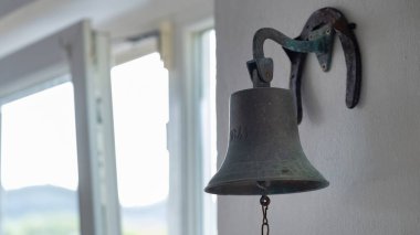 Beyaz bir duvara monte edilmiş klasik metal bir çan, kırsal bir at nalı desteği ve zinciri var. Arka plan yumuşak doğal ışığı ve açık pencereleri gösteriyor. Gelenek, konukseverlik, deniz dekoru veya kırsal yaşam ile ilgili konular için ideal.