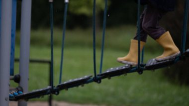 Sarı lastik çizme giyen bir çocuğun oyun parkında ip köprüden geçerken yakın plan görüntüsü. Açık hava oyunları, denge becerileri, oyun alanı güvenliği ve çocukların fiziksel gelişimi için mükemmel.