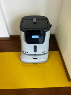 Köşedeki duvarın yanında sarı ve kahverengi bir zemine yerleştirilmiş otonom servis robotu. Küçük beyaz robotun ön ekran ekranı, sensörleri ve kompartımanları var.