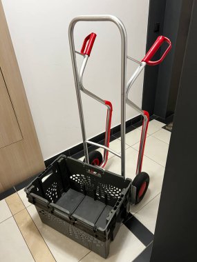 Kırmızı saplı metal el arabası ve kapalı bir koridorda fayanslı zemine konmuş katlanabilir siyah plastik sandık. Teslimat, ulaşım, taşınma, depolama ve