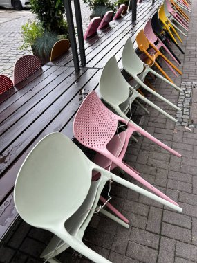 Yağmurdan sonra ıslak bir kaldırımda tahta sıralara yaslanmış renkli plastik sandalyeler. Pastel ve parlak renkler açık hava kafeleri ve sokak kafeleri için uygun canlı bir kontrast oluşturuyor.