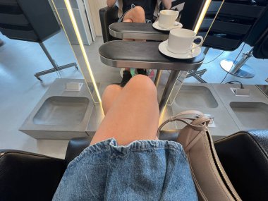 Salon sandalyesinden bir kişinin, aynanın önündeki küçük bir masaya yerleştirilmiş kahve fincanlarıyla rahatladığını gösteren bir görüntü. Fotoğraf, bir kuaför salonunun günlük atmosferini, güzellik tedavi molalarını ve