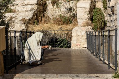 A Jewish man covered in a tallit or prayer shawl holds a silver cup while seeking financial donations near the Western Wall in Jerusalem, Israel.  clipart