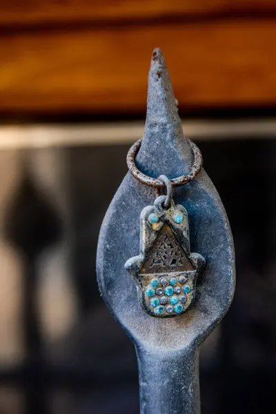 Küçük bir hamsa muskası. Hem Yahudi hem de İslam kültürlerinde korunmayı temsil eden sembolik bir el. Turkuaz boncuklarla süslenmiş ve metal bir çit direğine asılmış..