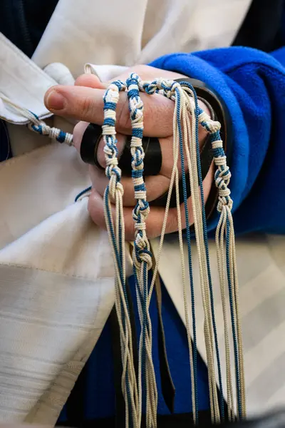 Closeup of the hand of an Orthodox, Jewish man holding blue techelet and white tzitzit strings while reciting the Shema Yisrael prayer during Jewish morning prayers.	