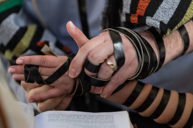 Başka bir yetişkinin yardımıyla, bir bar mitzvah çocuğu tefillininin siyah deri kayışlarını koluna, eline ve parmaklarına sarıp Yahudi sabah namazına hazırlanıyor.. 