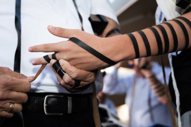 Başka bir yetişkinin yardımıyla, bir bar mitzvah çocuğu tefillininin siyah deri kayışlarını koluna, eline ve parmaklarına sarıp Yahudi sabah namazına hazırlanıyor.. 