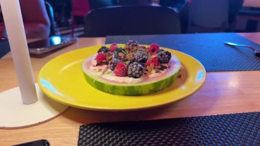 Meyve tabağı ilginç meyveli sandviç karpuzlu yoğurt kremalı karpuz dondurulmuş böğürtlen füme ahududu böğürtlenli mikroyeşillikler sarı tabak ile restorandaki ahşap masada. Yüksek kalite