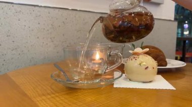 Yeşil bluz ve beyaz önlük giymiş ev hanımı huzurlu bir sabah boyunca çiçekli, pastalı ve klasik kitaplı rahat mutfak masasında şeffaf cam bardağa bitki çayı döküyor. 