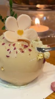 Kremalı, bej bir tatlı kaşıkla kesilmiş, kremalı tatlı. Badem dacquoise, ahududu karışımı, karamelli fındıklı çıtır tabaka ve ahududu tozu, acı çikolataya dayalı krema..