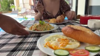 Güneşli bir günde otelde kahvaltı yapan bir kadın. Mavi yüzme havuzunun yanındaki villada sabah yemeği. Havuz başında yiyecek dolu bir masa, sağlıklı kahvaltı ve tatil beldeleri için meyve tabağı. Tropikal lüks yaşam tarzı