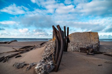 Çökmüş bir kaleden gelen paslı metal parçaları ile deniz kıyısı, çarpıcı bir gökyüzüne karşı güzel bulutlarla, tarihi ve doğayı güçlü bir kıyı sahnesinde harmanlıyor.
