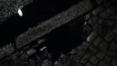 Islak bir kaldırım taşı sokağının su birikintisinde yansımaları olan soyut bir gece görüntüsü. Karanlık, karamsar atmosfer ve dokular görsel olarak ilgi çekici bir şehir manzarası yaratıyor..