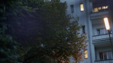 Berlin 'de alacakaranlık sahnesi, ışıklı pencereli bir apartman dairesi. Ön planda bir sokak lambası ve bir ağaç derinlik ekler. Tipik bir şehir akşam atmosferini yakalar.