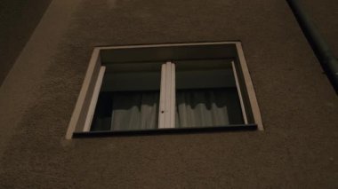 Geceleri bir apartmanın penceresine yakın çekim, perdeleri açık. Düşük ışık ve basit kompozisyon binanın dış yapısını vurgular..